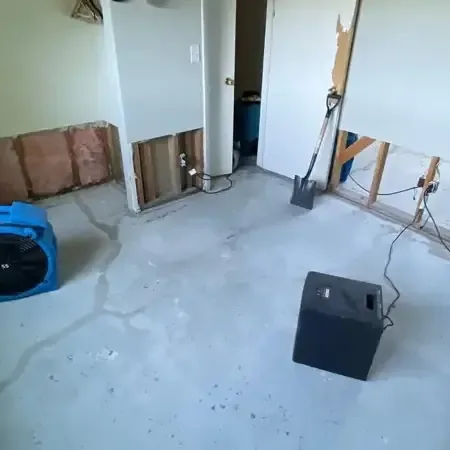 Air mover and dehumidifier on a bare concrete floor beside cut out lower drywall